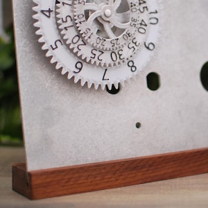 May include: A white metal clock with a circular face and a rotating gear mechanism. The clock face has numbers from 1 to 12 and markings for minutes. The clock is on a wooden base.