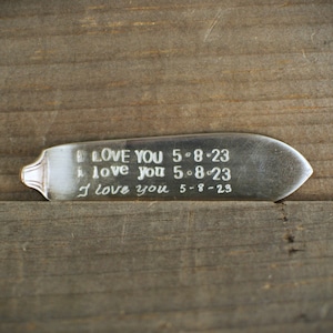 Op de afbeelding: Een zilveren metalen bladwijzer met de inscriptie "I love you 5-8-23" die drie keer wordt herhaald.