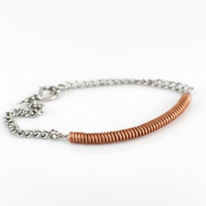 Op de afbeelding: Een zilveren kettingarmband met een koperkleurig spoelontwerp. De armband heeft een tuimelsluiting en een contrasterende mix van materialen. De koperen spoel is een prominent designelement.