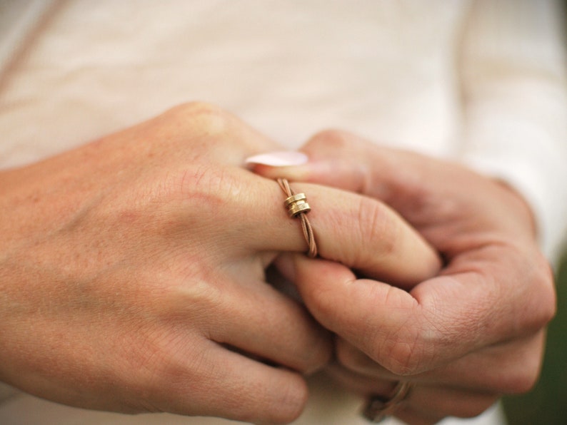 Pode incluir: Um close-up de uma m&atilde;o usando um anel de couro marrom com dois detalhes de metal dourado.