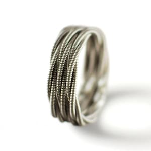 Op de afbeelding: Een zilveren ring gemaakt van opgerolde metalen draad, waarschijnlijk gitaarsnaren, tegen een witte achtergrond. De ring heeft een gedetailleerde textuur met zichtbare groeven en een metallic glans. Het ontwerp is eenvoudig maar uniek, en vormt een cirkelvormige band.