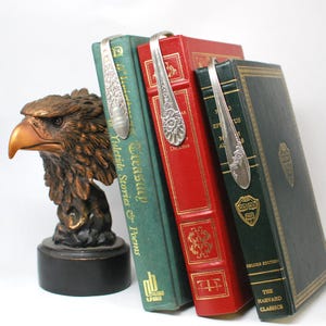 May include: A bronze eagle head bookend supports three vintage books with spoon bookmarks. The books are green, red, and green, with gold lettering. The spoon bookmarks are silver and have floral designs.