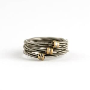 Op de afbeelding: Een handgemaakte ring van opgerolde, zilverkleurige metalen snaren. De ring is versierd met drie kleine, goudkleurige cilindrische kralen. Het ontwerp komt goed uit tegen een effen witte achtergrond.