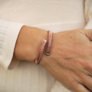 May include: A rose gold colored metal bracelet with a flexible, segmented design. The bracelet is worn on a person's wrist.
