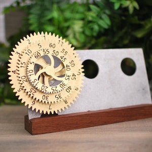 Puede incluir: Un soporte de madera con un mecanismo de engranaje de color dorado. El mecanismo de engranaje tiene números del 1 al 60. El soporte es de madera y tiene un acabado marrón oscuro.
