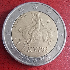May include: A silver and gold 2 Euro coin featuring a woman riding a bull with the text "ΕΥΡΩΠΗ" and "2 ΕΥΡΩ" around the edge.