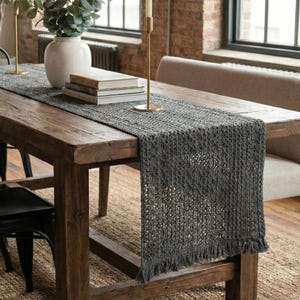 May include: A rustic wooden dining table is set with a gray woven table runner. A white vase with greenery, books, and gold candle holders with lit candles are on the table. Black chairs and a bench are around the table.