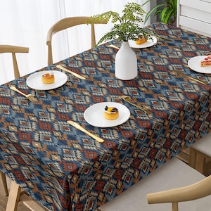 Può includere: Una tovaglia rettangolare con un motivo geometrico nei toni del blu, marrone e beige. La tavola è apparecchiata con piatti bianchi, posate dorate e un vaso bianco con verde.