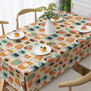 May include: Cream-coloured rectangular tablecloth featuring a retro geometric design in teal, orange, pink, and black. The tablecloth is set on a wooden table with white plates, gold cutlery, and a white vase with greenery.