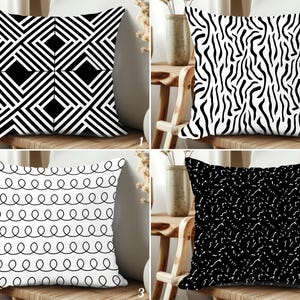 Könnte beinhalten: Vier dekorative Zierkissen mit geometrischen Mustern. Die Kissen weisen schwarz-weiße Designs auf, darunter ein Rautenmuster, Zebrastreifen, ein Schleifenlinienmuster und ein gesprenkeltes Design. Jedes Kissen ist quadratisch und liegt auf einem Holzstuhl.