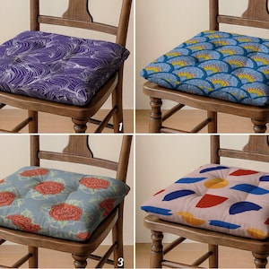 May include: Four square chair cushions with various patterns. One features a purple and white wave design, another has a blue and yellow fan pattern, a third has red roses on a grey background, and the last has blue, red, and yellow shapes.