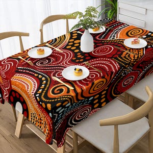 Puede incluir: Un mantel rectangular con un diseño abstracto vibrante en rojo, naranja, negro y blanco. El patrón presenta formas arremolinadas y líneas punteadas. La mesa está puesta con platos blancos, tenedores dorados y pequeños postres.