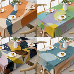 May include: Four colorful tablecloths with abstract designs are displayed on wooden tables. Each tablecloth features a unique pattern of overlapping shapes in various colors, including orange, green, purple, and blue. Each table is set with white plates, gold cutlery, and a small vase with greenery.