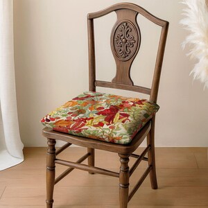 A wooden chair with a colorful floral cushion on the seat, placed on a wooden floor against a white wall.
