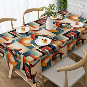 May include: A rectangular tablecloth with a geometric pattern in orange, teal, red, and beige. The design features overlapping shapes and lines. The table is set with white plates, gold cutlery, and a vase with greenery.
