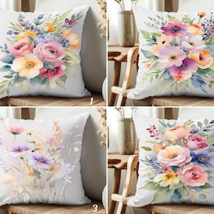 Op de afbeelding: Vier witte sierkussens met aquarel bloemmotieven. Elk kussen heeft een ander boeket bloemen in roze, paarse, gele en blauwe tinten, met groene bladeren. De kussens worden op een houten stoel getoond.