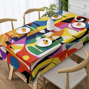 Pode incluir: Uma toalha de mesa retangular com um design abstrato vibrante em vermelho, amarelo, azul e verde. A toalha de mesa está colocada sobre uma mesa de madeira com cadeiras a condizer. Pratos brancos, talheres dourados e um vaso branco com vegetação completam o cenário.