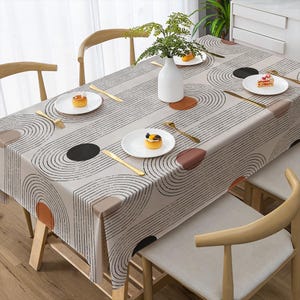May include: A beige rectangular tablecloth featuring a pattern of black curved lines and coloured circles. The table is set with white plates, gold cutlery, and a white vase with greenery. Wooden chairs surround the table.