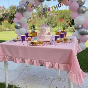May include: A pink birthday party table setting with a ruffled tablecloth, cake, cupcakes, and balloons. The cake is decorated with a tiara and heart-shaped designs. A banner reads "Happy Birthday."