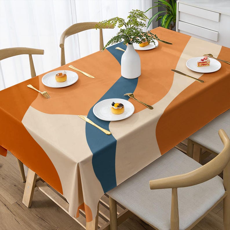 May include: A rectangular tablecloth with an abstract design in shades of orange, beige, and blue. The tablecloth is set on a wooden table with white plates, gold cutlery, and a white vase with greenery. The overall theme is home decor.