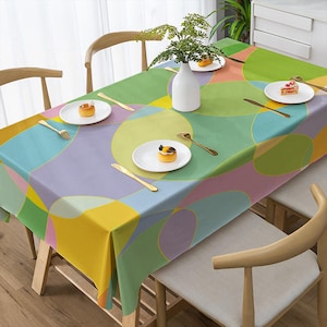 May include: A rectangular tablecloth with a colorful geometric pattern of overlapping circles in shades of yellow, green, blue, and pink. The table is set with white plates, gold cutlery, and a white vase with greenery.