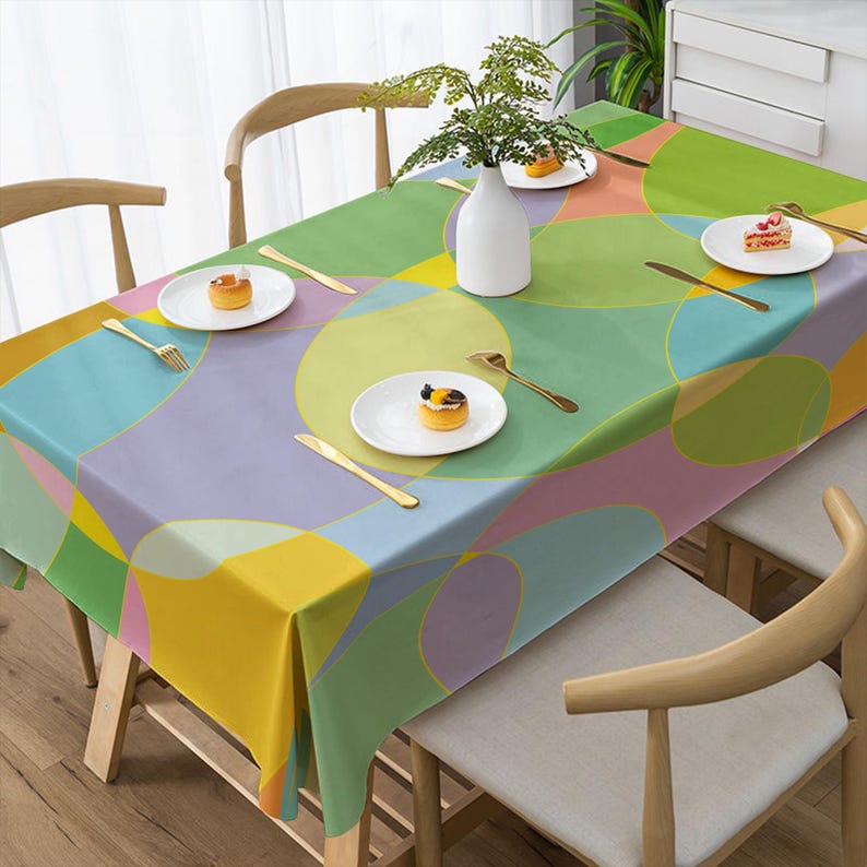 May include: A rectangular tablecloth with a colorful geometric pattern of overlapping circles in shades of yellow, green, blue, and pink. The table is set with white plates, gold cutlery, and a white vase with greenery.
