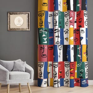 May include: Abstract art curtain featuring a repeating pattern of faces in a variety of colours, including yellow, red, blue, and green. The curtain hangs in front of a grey wall, next to a framed artwork and a grey armchair.