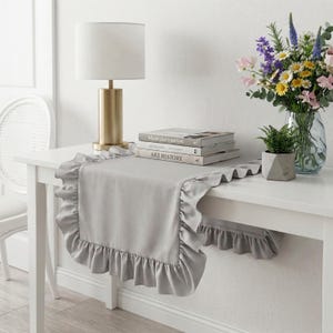 Pode incluir: Um caminho de mesa cinza-claro com uma borda franzida, colocado sobre uma mesa branca. O caminho está posicionado sob uma pilha de três livros, um candeeiro de tom latão com cúpula branca e um vaso de vidro com flores coloridas.