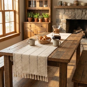 Pode incluir: Uma mesa de jantar rústica de madeira com um caminho de mesa creme com um padrão texturizado e borlas. A mesa está posta com canecas, uma cesta de croissants e uma cafeteira, criando uma atmosfera acolhedora e convidativa.