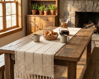 May include: A rustic wooden dining table with a cream-colored table runner featuring a textured pattern and tassels. The table is set with mugs, a basket of croissants, and a coffee maker, creating a cozy, inviting atmosphere.