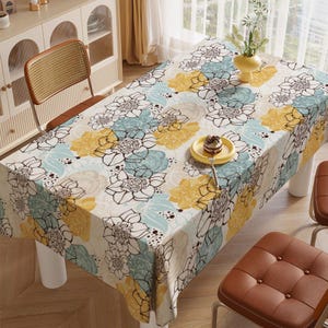 Op de afbeelding: Een rechthoekig tafelkleed met een bloemenpatroon in geel, blauw en wit bedekt een tafel. De bloemen zijn zwart omlijnd. Een klein geel bord met een dessert en een vork staat op het tafelkleed. Twee bruine stoelen zijn zichtbaar.