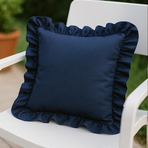 May include: A square navy blue pillow with a ruffled edge. The pillow is made of a soft fabric and is sitting on a white chair. The ruffled edge adds a decorative touch to the pillow.