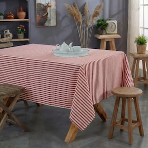 Puede incluir: Un mantel a rayas rojas y blancas cubre una mesa de madera. Un juego de té blanco descansa sobre una bandeja decorativa. La escena incluye taburetes de madera, una mesa pequeña y objetos decorativos.