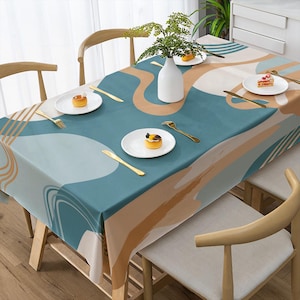 May include: A rectangular tablecloth with an abstract design in shades of teal, beige, and white. The table is set with white plates, gold cutlery, and a white vase with greenery. Wooden chairs surround the table.
