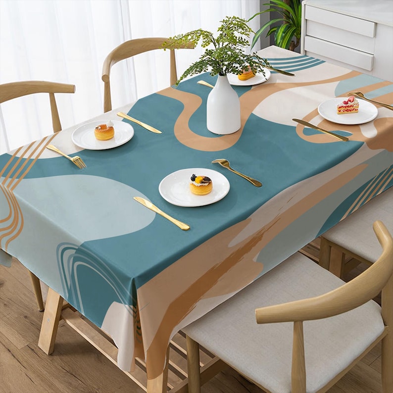 May include: A rectangular tablecloth with an abstract design in shades of teal, beige, and white. The table is set with white plates, gold cutlery, and a white vase with greenery. Wooden chairs surround the table.