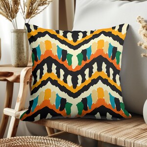 Puede incluir: Cojín decorativo cuadrado con un diseño abstracto vibrante. El patrón presenta líneas en zigzag negras con pinceladas de naranja, amarillo, azul y verde sobre un fondo crema. El cojín está colocado sobre una silla de madera.