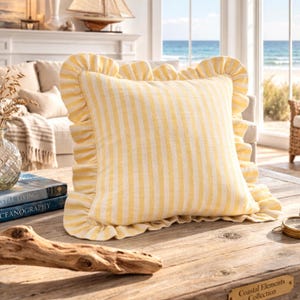 Op de afbeelding: Een vierkant decoratief kussen met gele en witte verticale strepen en een gerimpelde rand. Het kussen ligt op een houten tafel met een kustthema, met uitzicht op de oceaan op de achtergrond.