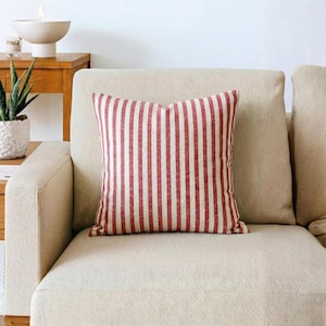 Könnte beinhalten: Ein quadratisches Zierkissen mit rot-weißem Streifenmuster. Das Kissen liegt auf einem beigefarbenen Sofa. Es ist ein Einrichtungsgegenstand.