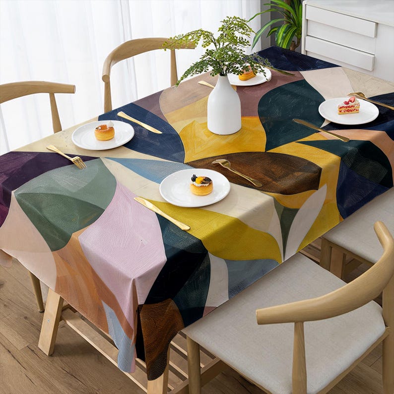 May include: A colorful abstract patterned tablecloth with a geometric design in shades of yellow, blue, pink, and brown. The table is set with white plates, gold cutlery, and a white vase with greenery. The table is surrounded by wooden chairs.