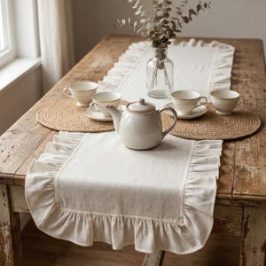 May include: A cream-coloured, ruffled table runner adorns a rustic wooden table, alongside a teapot, teacups, and woven placemats. A clear glass vase with greenery adds a natural touch to the tea setting. The table has a distressed finish.