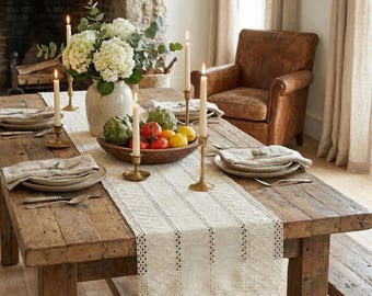 May include: A rustic wooden dining table set for a meal. The table is adorned with a cream-colored runner, candles in brass holders, plates, napkins, and a wooden bowl filled with fruit and artichokes. A vase of white flowers adds a touch of elegance.