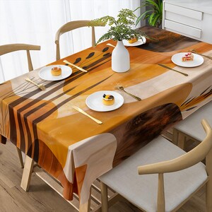 May include: A rectangular tablecloth with an abstract design in shades of orange, brown, and beige. The table is set with white plates, gold cutlery, and a white vase with greenery. The table is surrounded by wooden chairs.