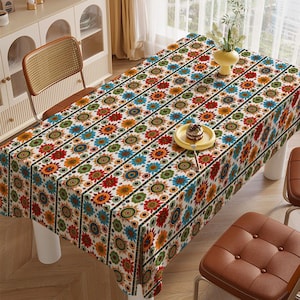 May include: Rectangular tablecloth with a vibrant floral pattern in red, orange, blue, and green on a white background. The design features repeating stylized flowers with black striped borders. A yellow plate with a small cake sits on the table.