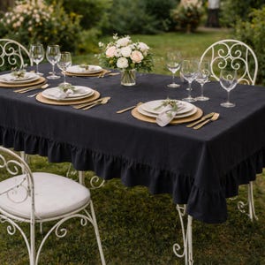 May include: An outdoor dining table set for six, featuring a dark blue linen tablecloth with a ruffled edge. Each place setting includes a white plate, gold cutlery, and a wine glass. A vase of flowers sits in the centre of the table.