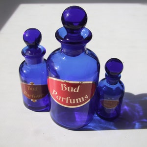 Pode incluir: Três garrafas de vidro azul cobalto com letras douradas em etiquetas vermelhas. A garrafa maior diz "Bud Parfums". As garrafas menores dizem "Bud Parfums".
