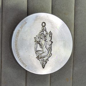 May include: A circular silver-toned metal stamp featuring an engraved design of a winged fairy figure in a seated position. The text KING'S TREASURES FAIRY_0011L is etched along the left edge of the flat, polished metallic surface of the tool.