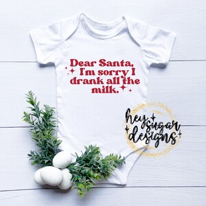 May include: White baby onesie with red text that reads "Dear Santa, I'm sorry I drank all the milk."  The text is surrounded by small gold glitter stars.  The bottom right corner of the onesie has a gold glitter circle with the text "hey sugar designs".