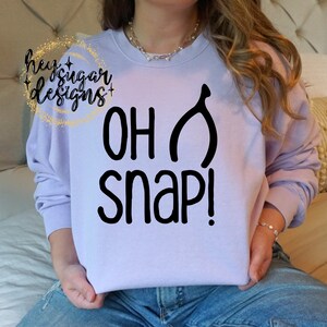 May include: A lavender sweatshirt with the text "OH SNAP!" and a black turkey leg graphic.