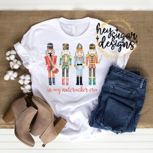 May include: White t-shirt with a graphic of four nutcracker soldiers in watercolor style. The text "in my nutcracker era" is printed below the soldiers. The shirt is laid out on a wooden surface with a pair of brown boots and blue jeans.