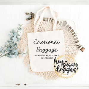 Op de afbeelding: Een witte tote bag met de tekst "Emotional Baggage" en de zin "Just kidding, did you really think it could fit in here?" erop gedrukt. De tas ligt op een wit oppervlak met een beige geweven wandkleed en groen gebladerte op de achtergrond. De tekst "heysugar designs+ " is in zwart met gouden glitteraccenten gedrukt.